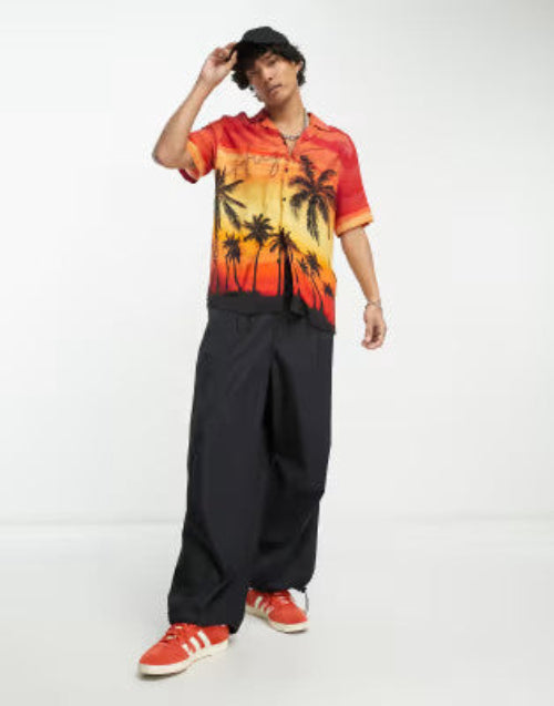 Translation missing: en.A model wearing a short-sleeve button-up shirt with a vibrant sunset and palm tree graphic, paired with loose black pants and red sneakers, set against a plain white studio background