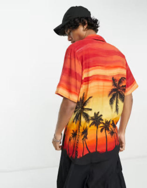 Translation missing: en.A man wearing a short-sleeve button-up shirt with a sunset and palm tree graphic, shown from the back. The shirt features a gradient of red, orange, and yellow with black palm tree silhouettes