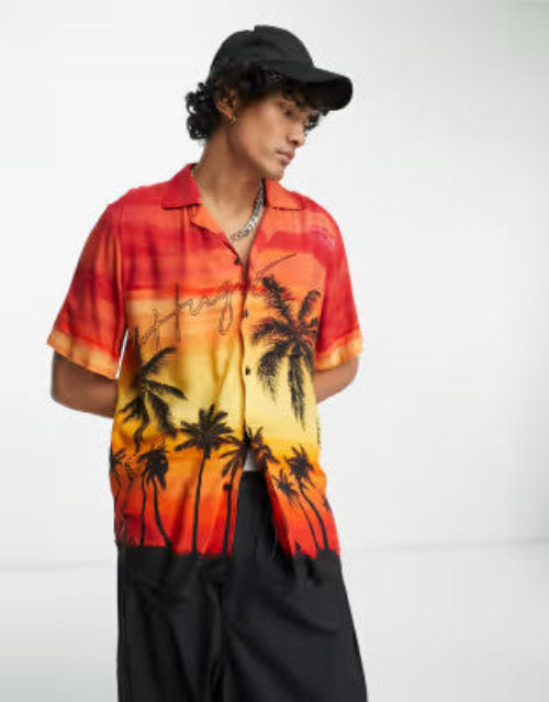 Translation missing: en.A man wearing a short-sleeve button-up shirt with a sunset gradient and black palm tree print, paired with black pants and a black baseball cap, photographed against a plain light gray studio background
